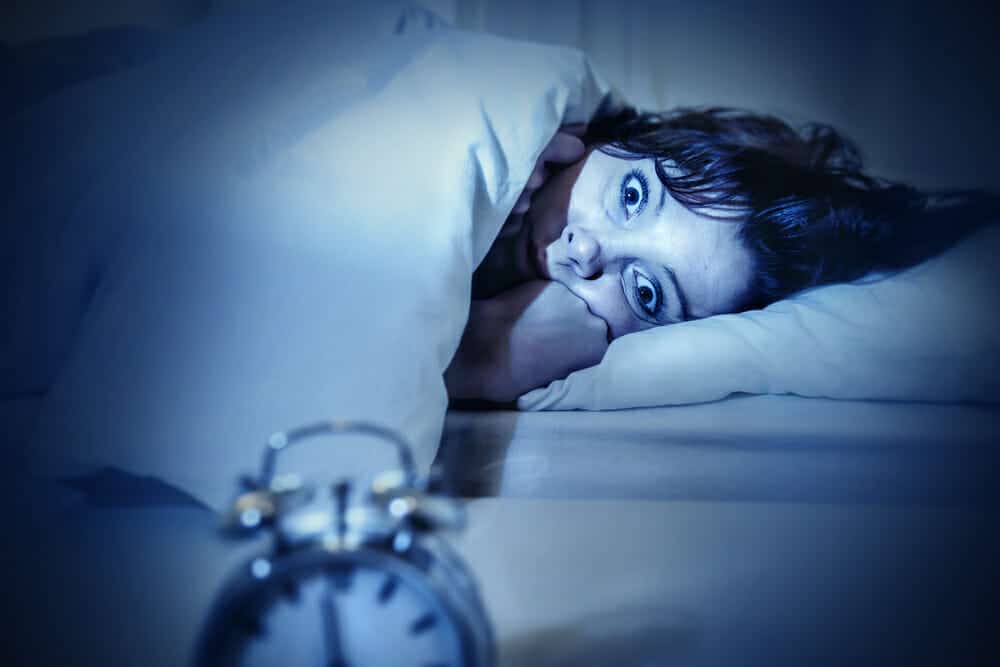 young woman in bed with alarm clock and eyes opened suffering insomnia and sleep disorder thinking about his problem on dark studio lighting in sleeping and nightmare issues