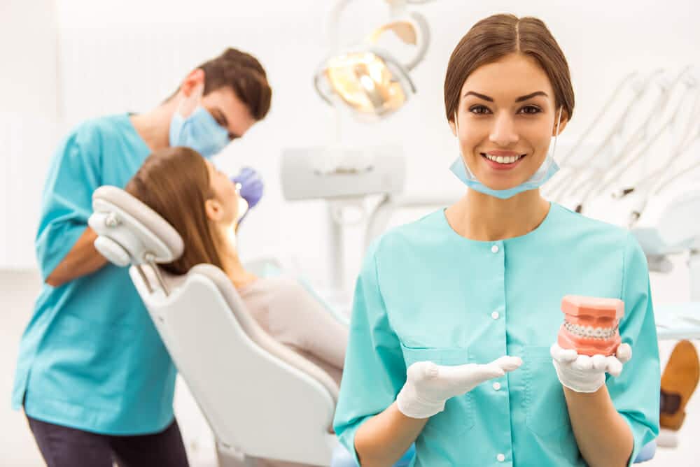Young assistant holding a plastic model teeth, in the background dentist treats a patient