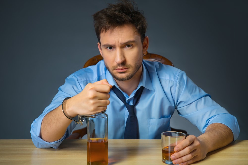 In thrall. Cheerless drunk man holding bottle of alcohol in handcuffs and sitting at the table while drinking