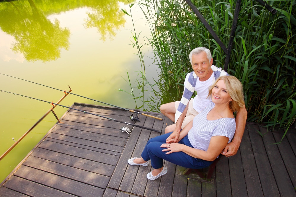 Senior couple relaxing at the lakeside. Smiling senior woman and man sitting on the pier and fishing.